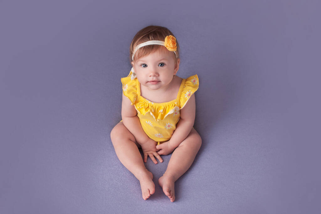 niña de un año en un traje de cuerpo amarillo se sienta sobre un fondo azul. - Foto, imagen