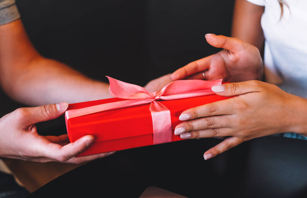 Genç adamın kız arkadaşına pembe kurdeleli kırmızı hediye kutusu verişi. Beklenmedik bir hediye alan kadın. Kutlama konsepti. Yüksek kalite fotoğraf - Fotoğraf, Görsel
