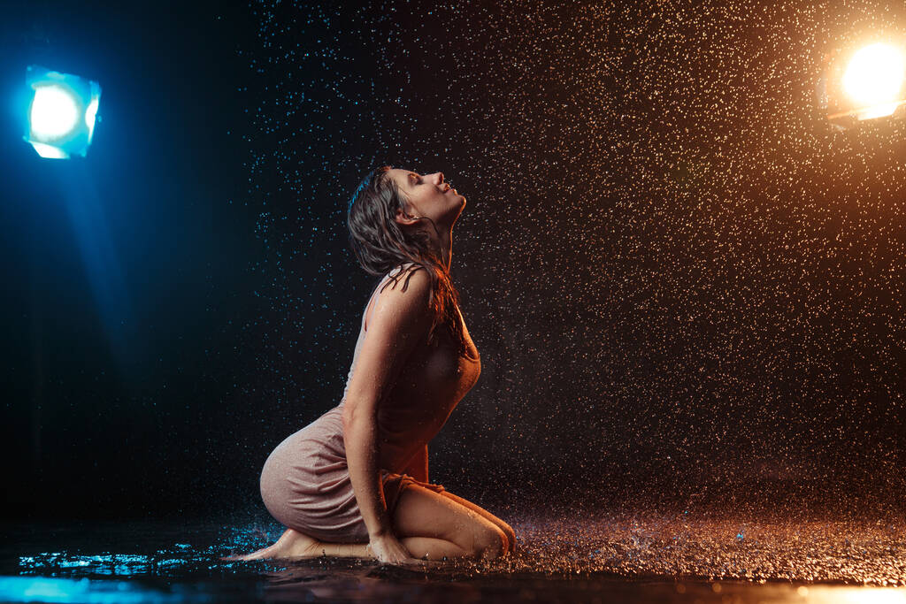 Красивая сексуальная девушка с капельками воды на лице в брызгах, освещенных оранжевым светом на темном фоне в студии. - Фото, изображение