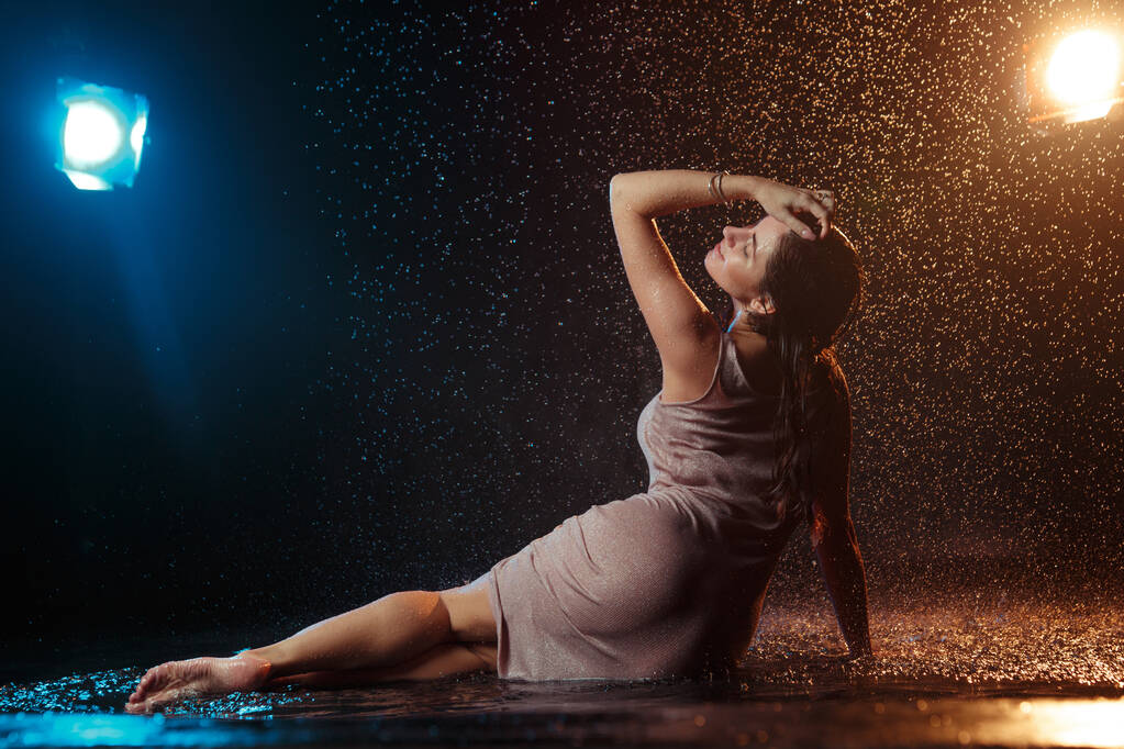 Menina sexy bonita com gotas de água em seu rosto em salpicos iluminados por uma luz laranja contra um fundo escuro no estúdio. - Foto, Imagem