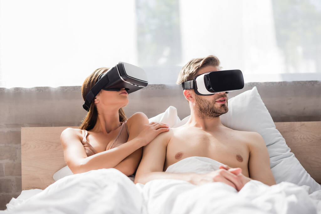 νέα γυναίκα αγγίζοντας shirless φίλο, ενώ χρησιμοποιώντας ακουστικά VR στο κρεβάτι - Φωτογραφία, εικόνα