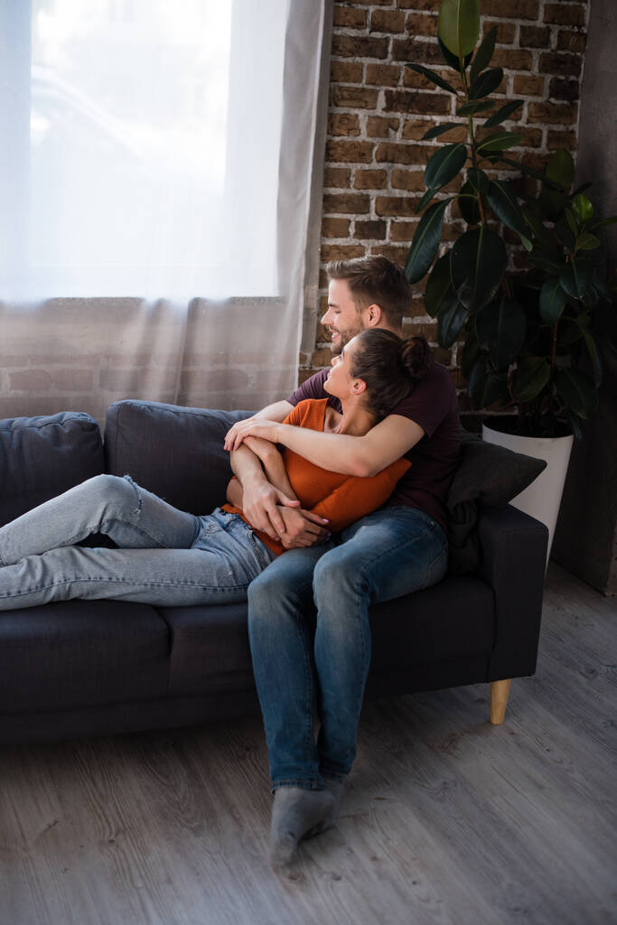 νεαρό ζευγάρι αγκαλιάζει ενώ αναπαύεται στον καναπέ στο σπίτι - Φωτογραφία, εικόνα