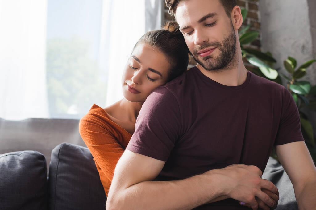 Zarte Frau mit geschlossenen Augen umarmt geliebten Mann von hinten, während sie auf dem Sofa sitzt - Foto, Bild