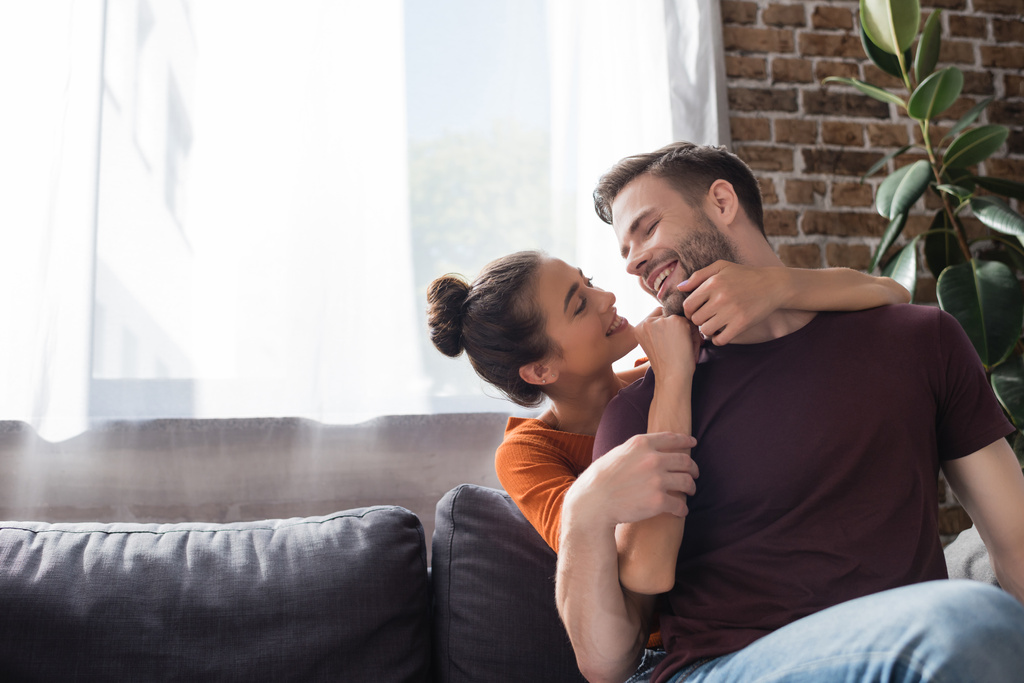 cheerful woman touching face of happy boyfriend while sitting on sofa at home - Photo, Image