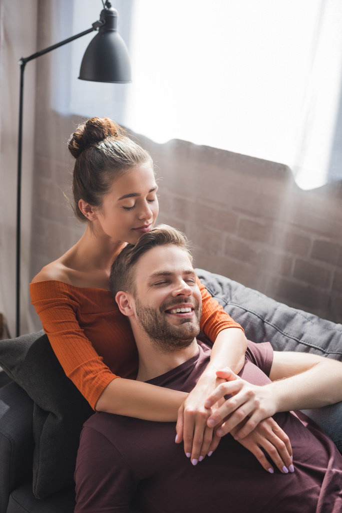 mulher feliz abraçando namorado sorridente enquanto descansa no sofá em casa - Foto, Imagem