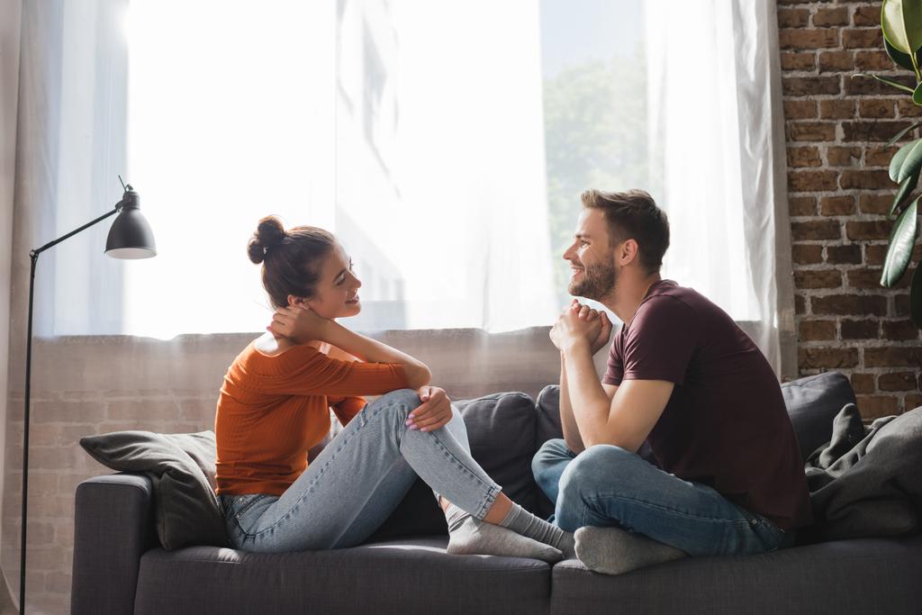 vista lateral do jovem casal sentado no sofá e falando enquanto olha um para o outro - Foto, Imagem