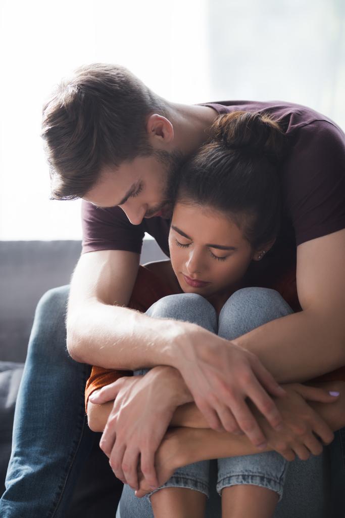 junger Mann umarmt und beruhigt geliebte Frau, die mit geschlossenen Augen auf Sofa sitzt - Foto, Bild