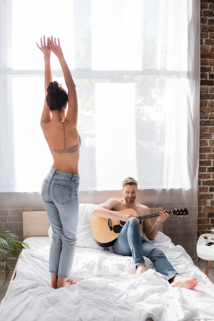 Вид сзади соблазнительной женщины, танцующей с поднятыми руками рядом с бойфрендом, играющим на гитаре в постели - Фото, изображение