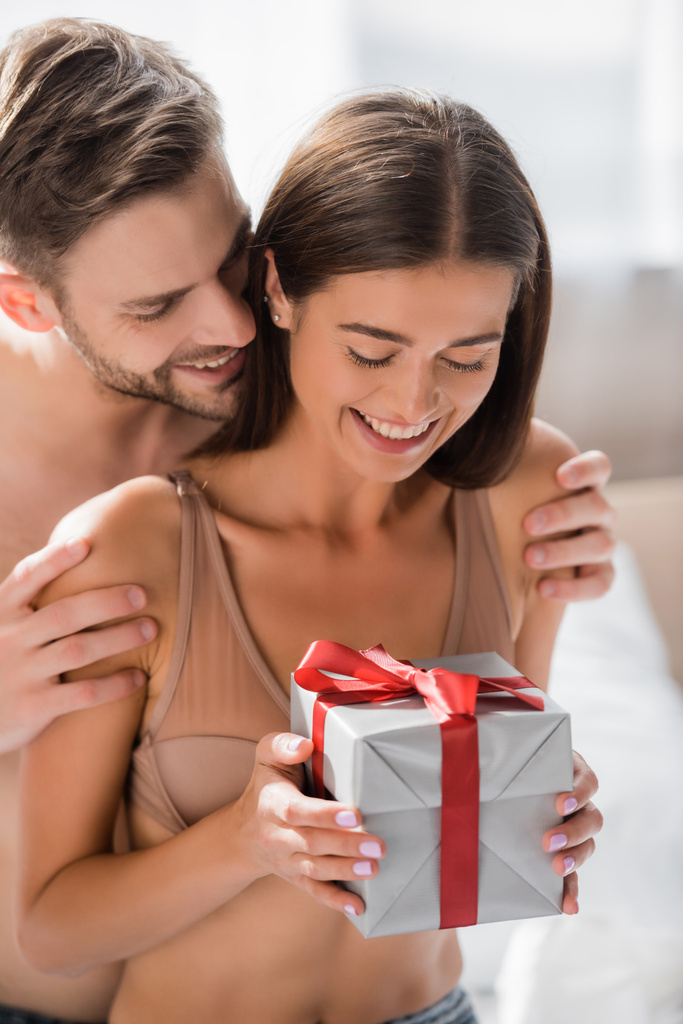 Улыбающийся мужчина обнимает счастливую девушку с подарочной коробкой в спальне - Фото, изображение