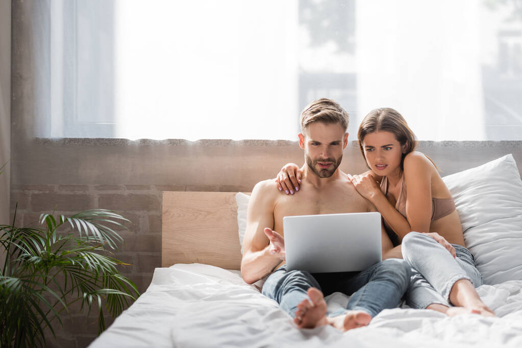 donna scioccata toccando fidanzato senza maglietta utilizzando il computer portatile a letto - Foto, immagini
