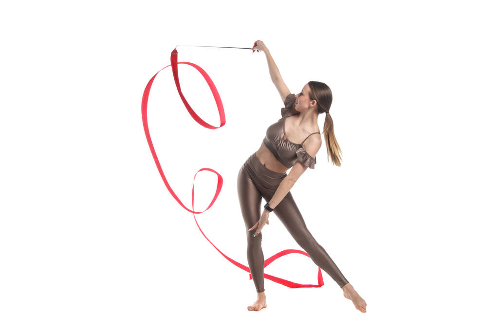 Стройная изящная гимнастка в бежевой спортивной одежде танцует с красной развивающейся лентой, изолированной на белом фоне. - Фото, изображение