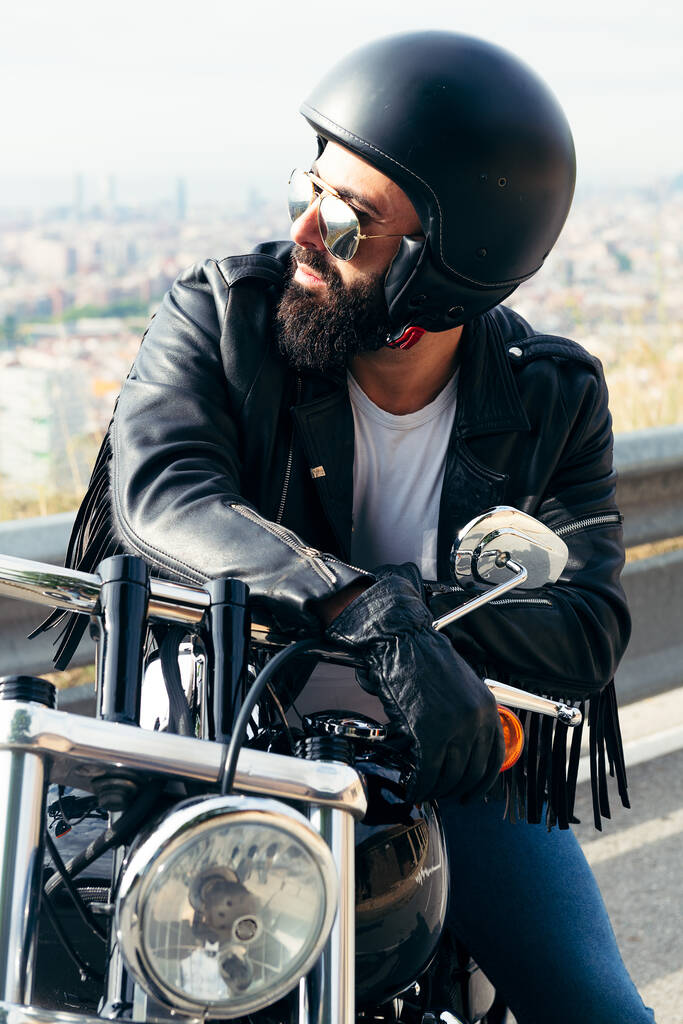 biker with helmet and leather jacket sitting on his motorbike with the city far away in the background, concept of freedom and rebel lifestyle - Photo, Image