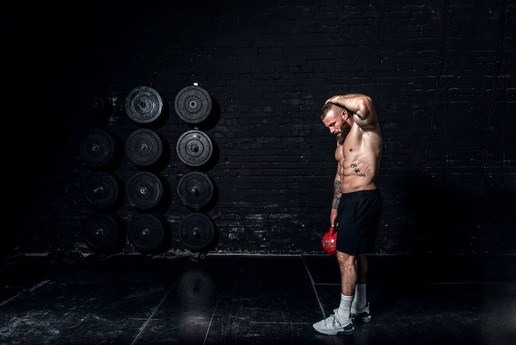 Νέοι ιδρωμένοι ισχυρή μυϊκή τακτοποίηση άνθρωπος με μεγάλους μυς κάνει side abs προπόνηση στο γυμναστήριο με το να στέκεται και τραβώντας προς τα πάνω βαρύ kettlebell με το ένα χέρι ως hardcore προπόνηση σταυρό - Φωτογραφία, εικόνα