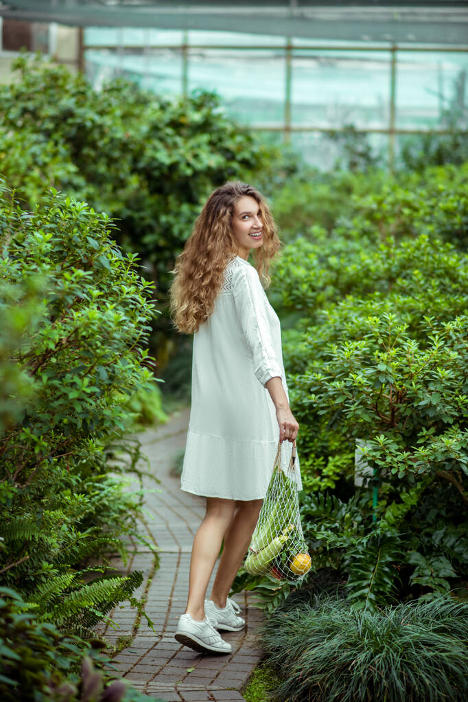 Frau in weißem Kleid trägt Tüten mit verschiedenen Gemüsesorten und fühlt sich wohl - Foto, Bild