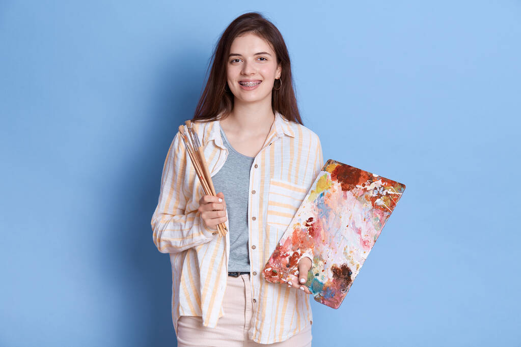 Attraktive junge kaukasische Künstlerin mit Malzubehör, Pinsel in der Hand, Farbmischung auf Palette, Dame mit weißem Hemd und weißer Hose. - Foto, Bild