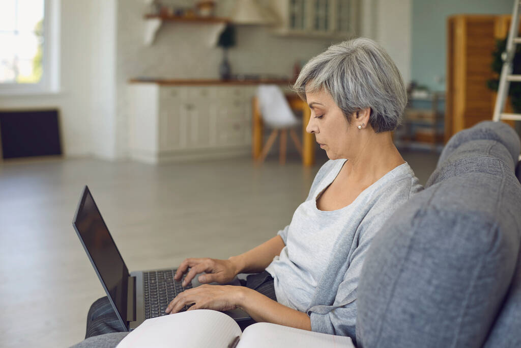 Vakava vanhempi nainen työskentelee kannettavan tietokoneen istuu iso nojatuoli kannettavan vieressä - Valokuva, kuva