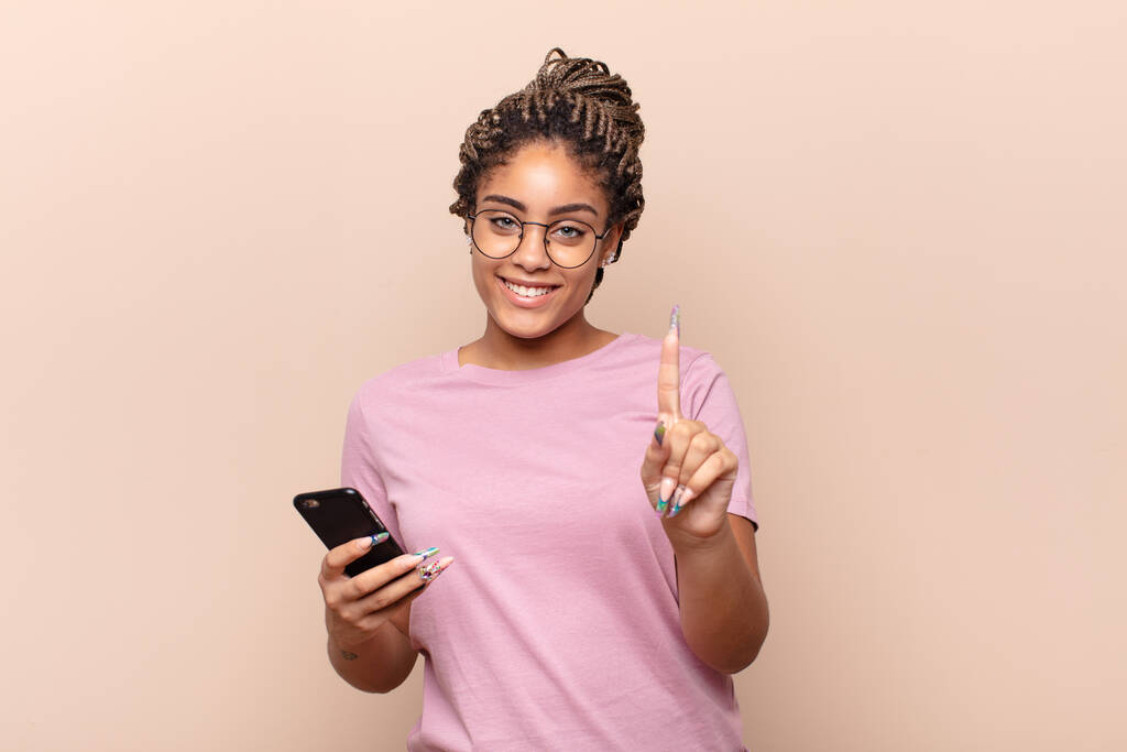 joven afro mujer sonriendo y buscando amigable, mostrando el número uno o primero con la mano hacia adelante, cuenta atrás. concepto de teléfono inteligente - Foto, imagen