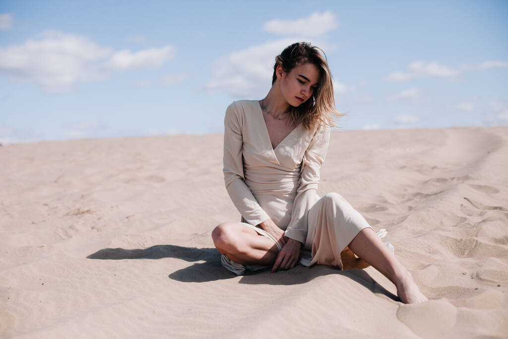 Ένα νέο, λεπτό κορίτσι με ένα μπεζ φόρεμα ποζάρει στον άνεμο στην έρημο. - Φωτογραφία, εικόνα