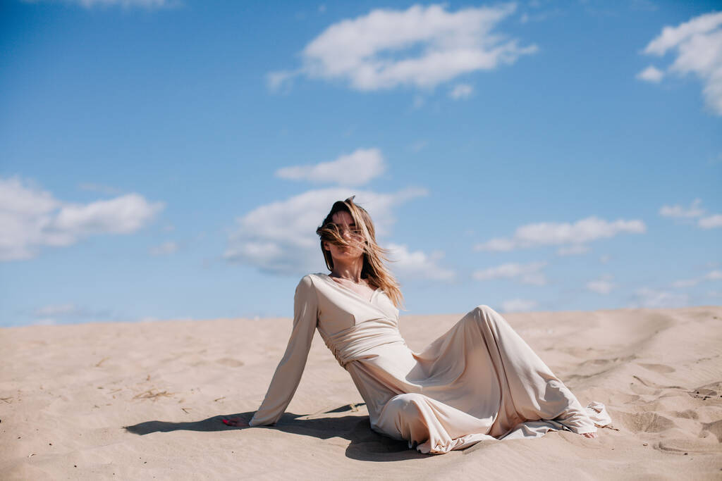 Młoda, szczupła dziewczyna w beżowej sukience pozuje na wietrze na pustyni - Zdjęcie, obraz