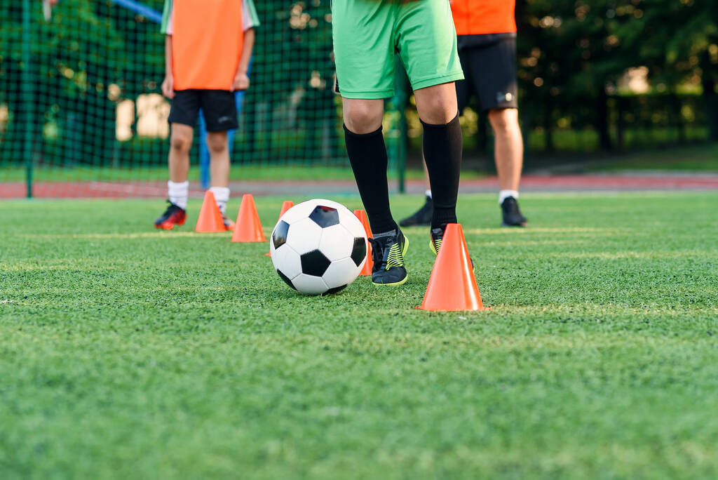 Close up menino em sportswear treina futebol no campo de futebol e aprende a circular a bola entre treinar cones laranja. - Foto, Imagem