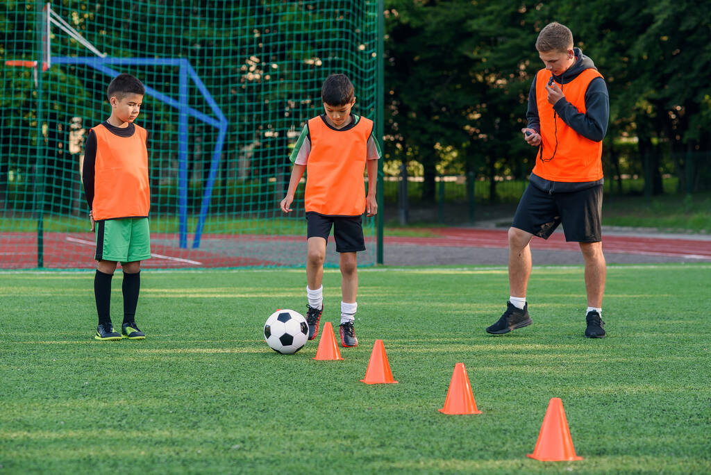 Hartnäckiger Teenager in Sportbekleidung trainiert Fußball auf dem Fußballplatz und lernt, den Ball zwischen Trainingskegeln zu kreisen. - Foto, Bild