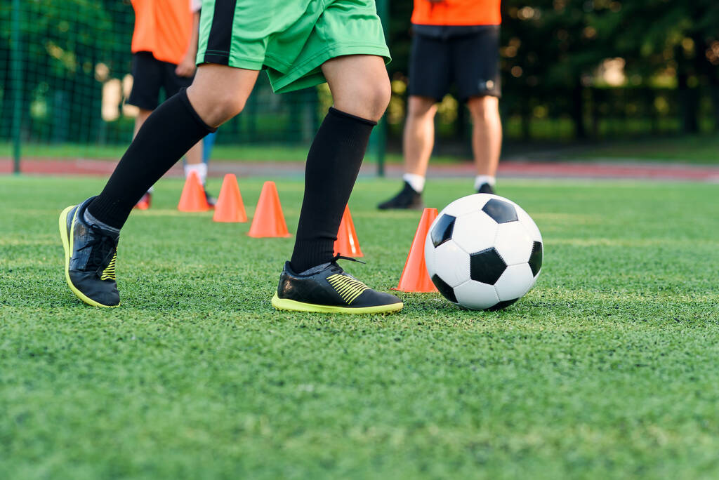 Настойчивый подросток в спортивной форме тренирует футбол на футбольном поле и учится кружить мяч между тренировочными конусами. - Фото, изображение