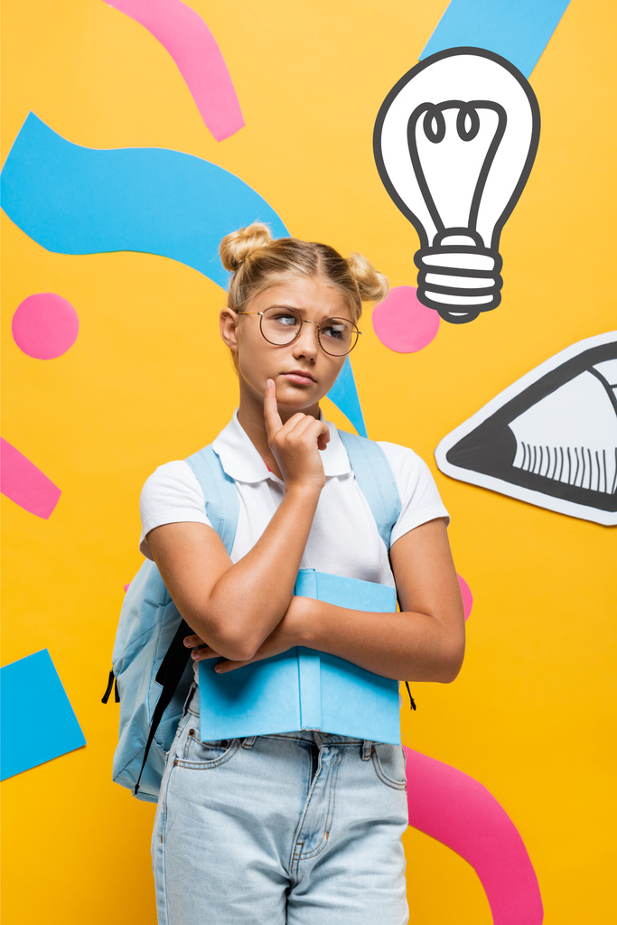 doordachte schoolmeisje in bril wegkijkend terwijl boek in de buurt van papier potlood, kleurrijke elementen en illustratie op geel - Foto, afbeelding