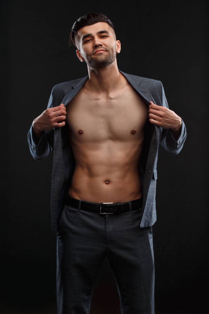 A young sexy man takes off his jacket worn over his naked body against a dark background. - Photo, Image