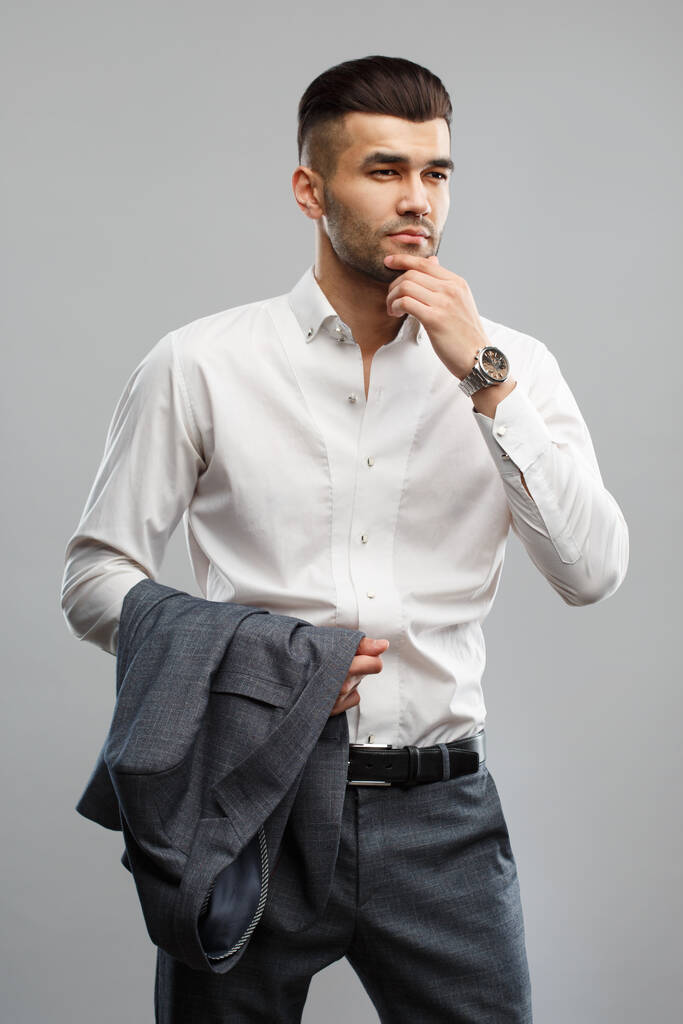 Joven hombre con estilo en una camisa blanca sostiene una chaqueta de traje en su mano aislado sobre un fondo gris. - Foto, imagen