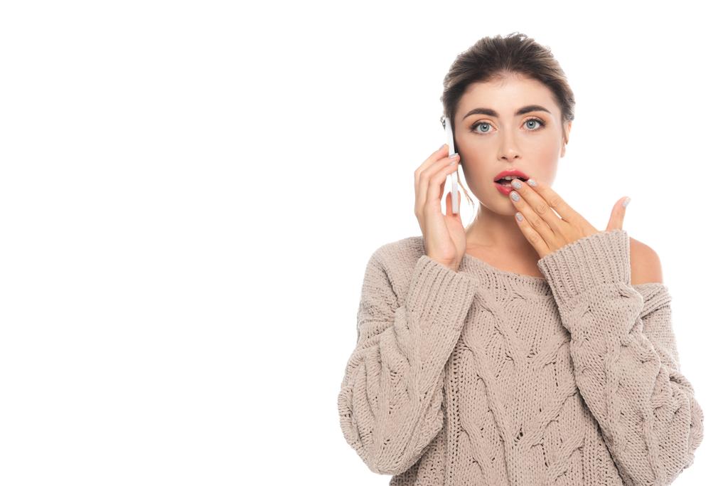 шокированная женщина в стильном свитере открытого цвета, закрывающем рот рукой, разговаривая на смартфоне, изолированном на белом - Фото, изображение