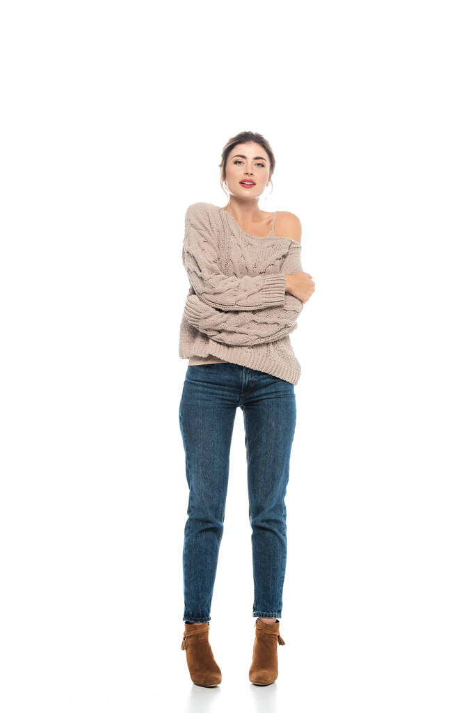 full length view of trendy woman in openwork sweater and jeans hugging herself while posing on white - Photo, Image