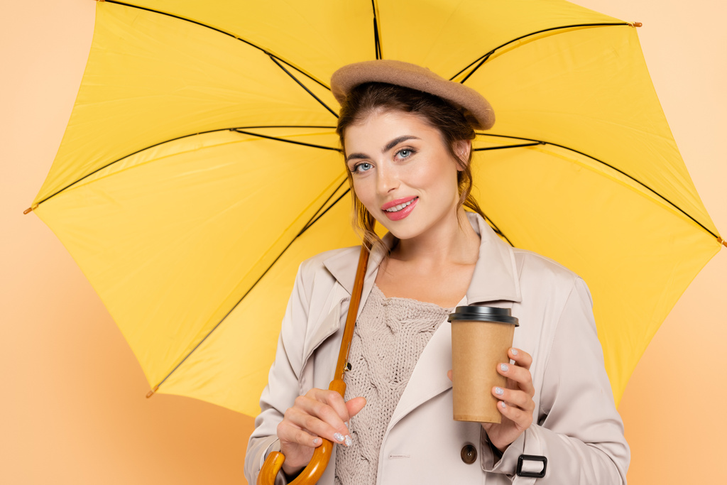 trendy vrouw in trench jas en baret houden koffie te gaan onder gele paraplu op perzik - Foto, afbeelding