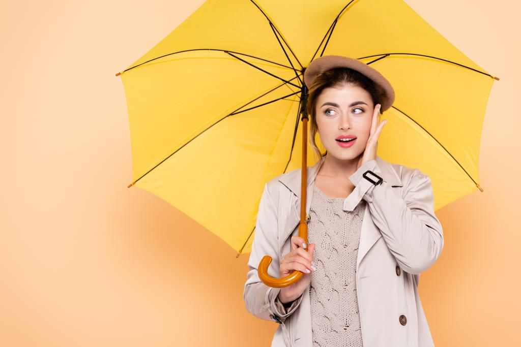 trendy woman in autumn outfit touching face and looking away under yellow umbrella on peach - Photo, Image