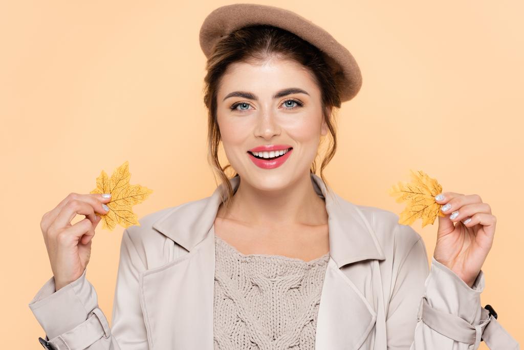 fashionable woman in autumn outfit holding yellow leaves while looking at camera isolated on peach - Photo, Image