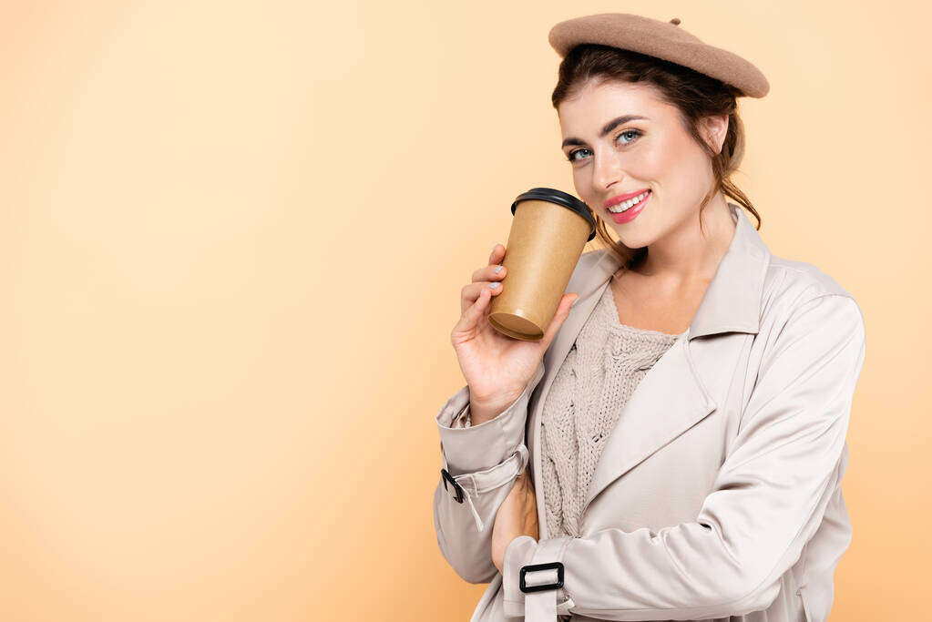 mladá žena v trenč kabát a baret drží kávu jít při pohledu na kameru izolované na broskev - Fotografie, Obrázek