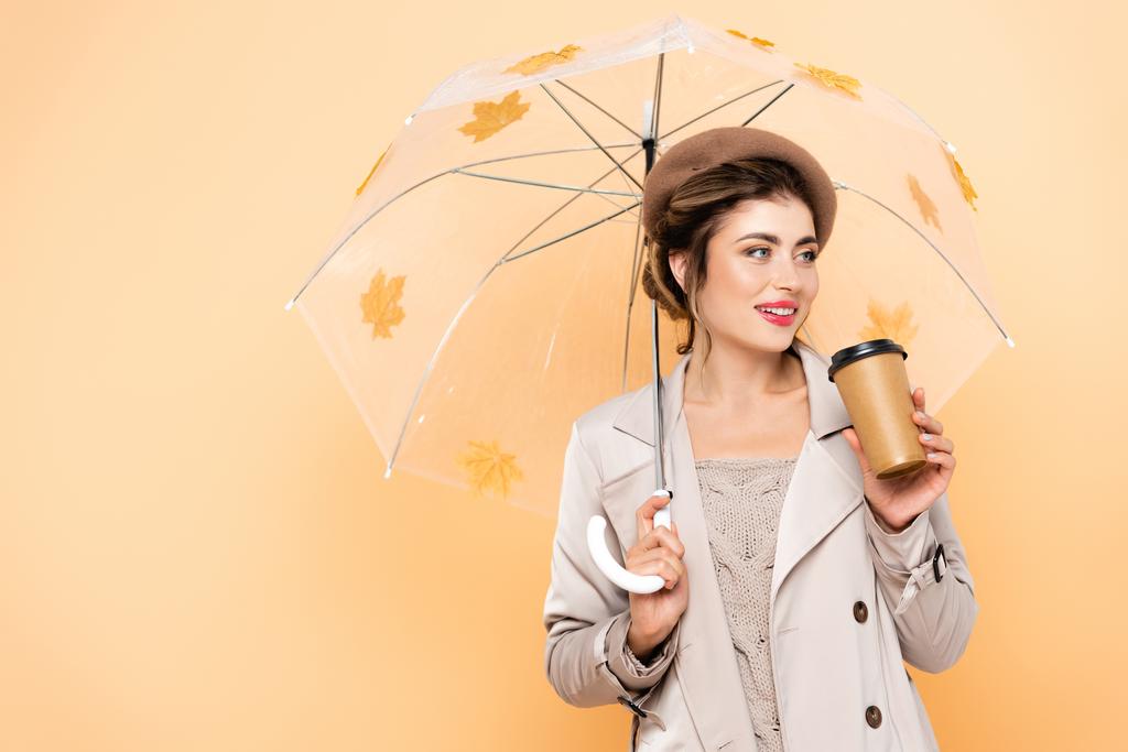 Trendfrau in Baskenmütze und Trenchcoat mit Kaffee zum Regenschirm und Herbstblättern auf Pfirsich - Foto, Bild