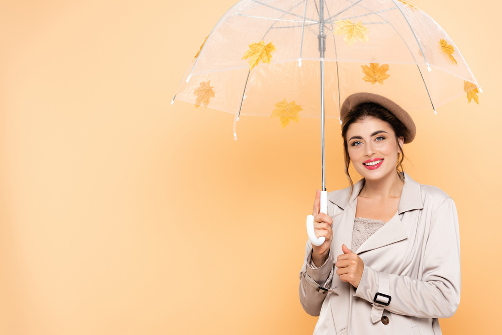 stylische Frau in Baskenmütze und Trenchcoat, die unter einem Regenschirm mit Herbstblättern auf Pfirsich in die Kamera blickt - Foto, Bild