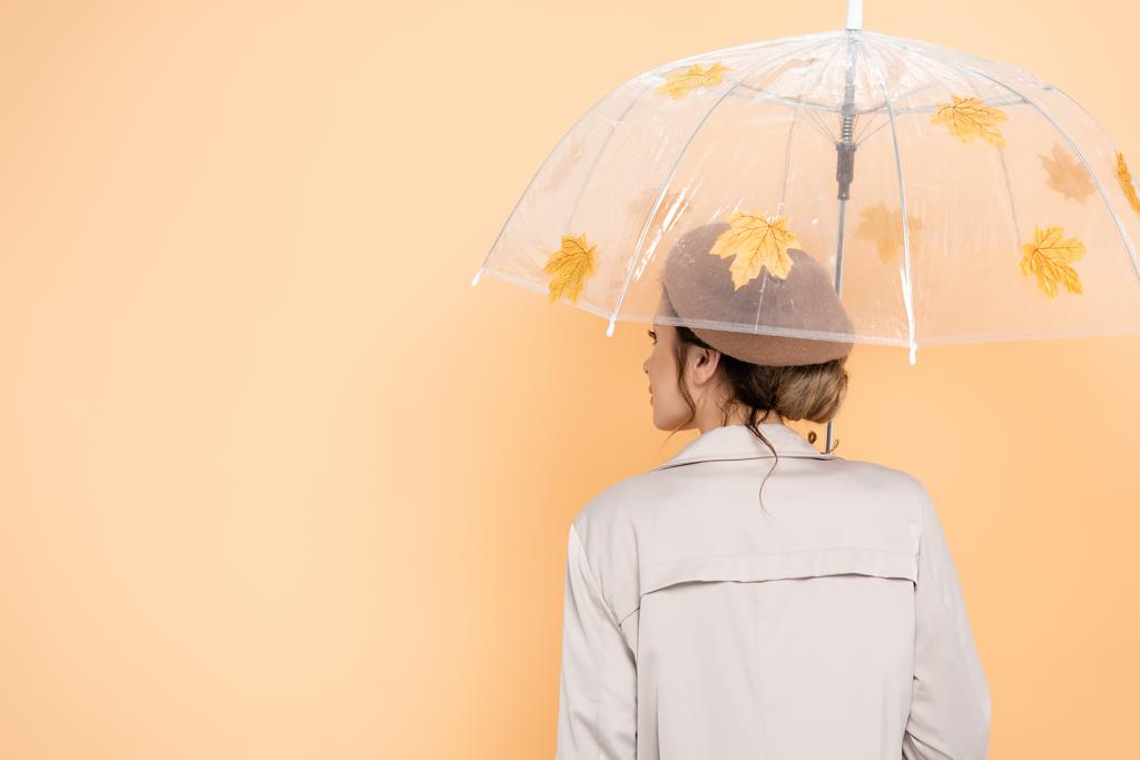 back view of trendy woman in beret and trench coat under umbrella, decorated with yellow leaves on peach - Photo, Image