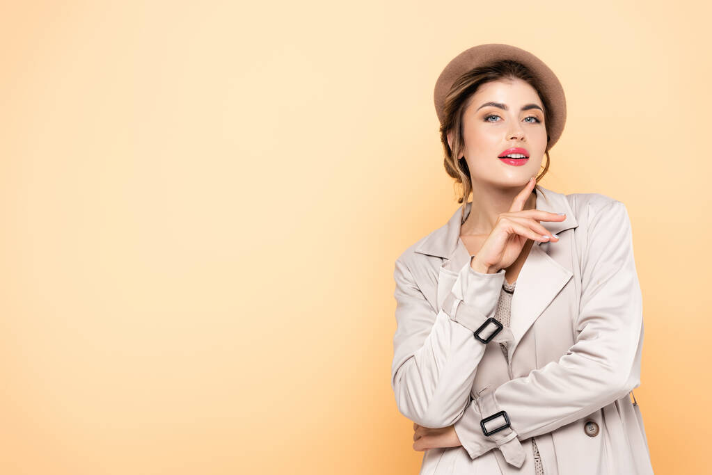 sensual woman in stylish trench coat and beret looking at camera on peach - Photo, Image