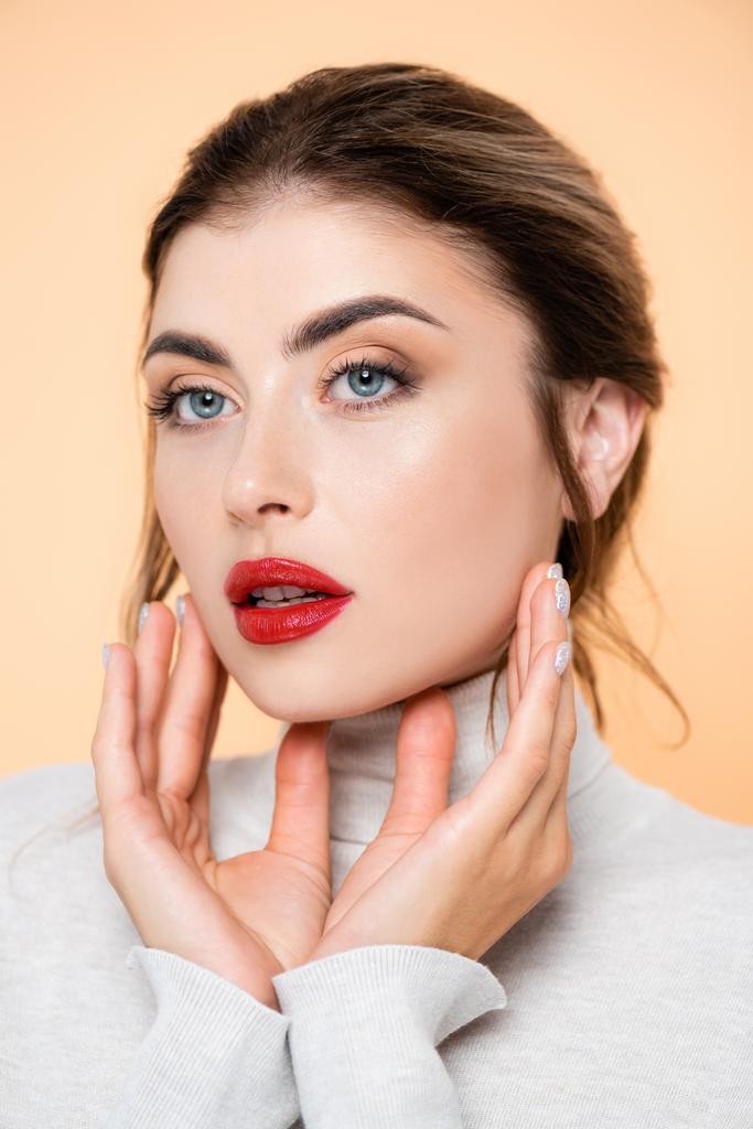 young woman with red lips looking away while touching face isolated on peach  - Photo, Image