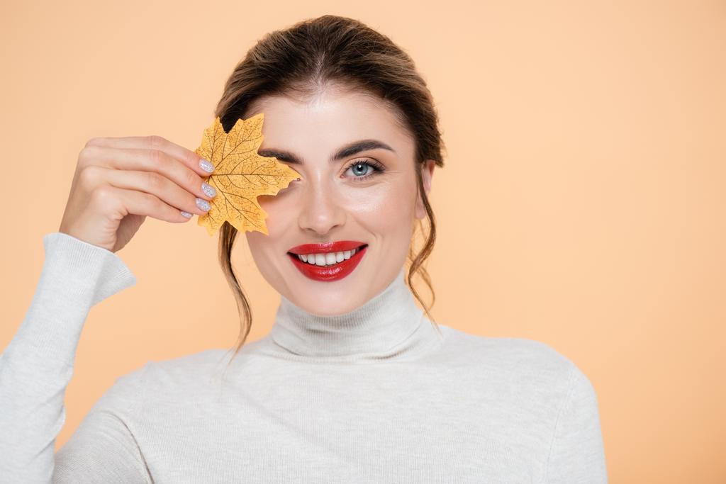 joyful woman with red lips looking at camera while covering eye with yellow leaf isolated on peach  - Photo, Image