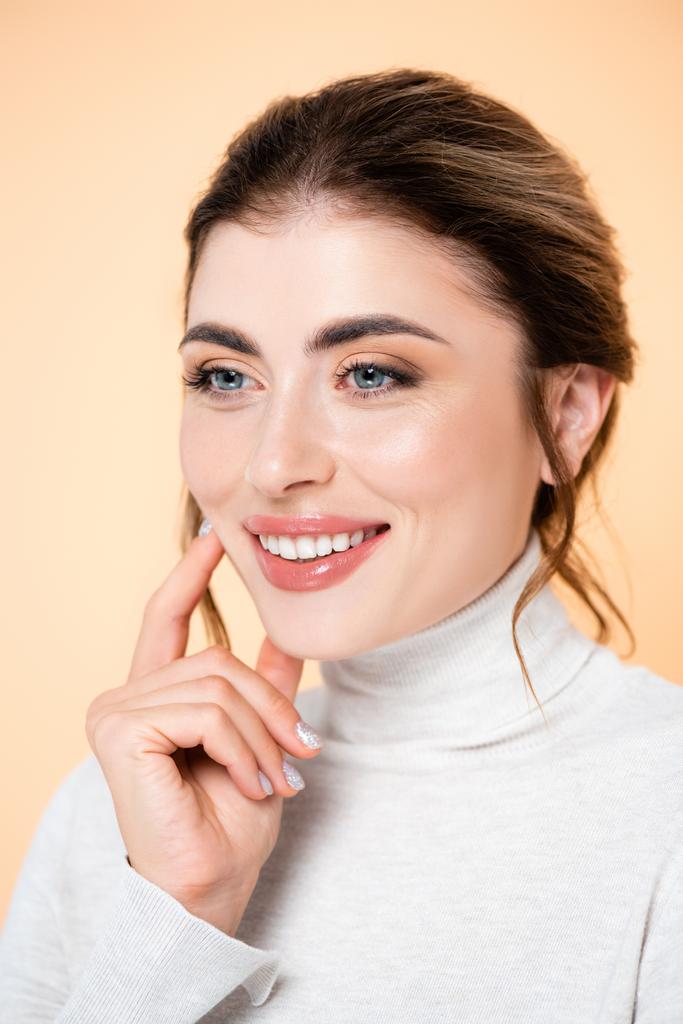 joyful woman in turtleneck touching face while looking at camera isolated on peach  - Photo, Image