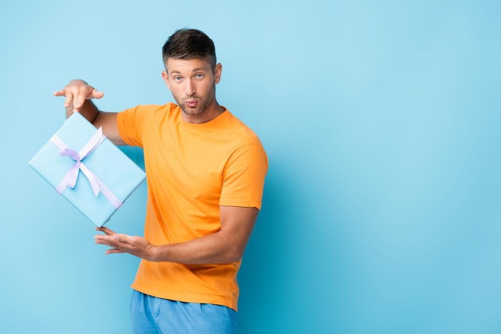 Mann im T-Shirt schmollende Lippen und hält verpackten Geschenkkarton auf blau - Foto, Bild