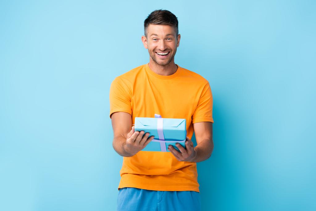 pleased man in t-shirt holding wrapped gift box on blue - Photo, Image