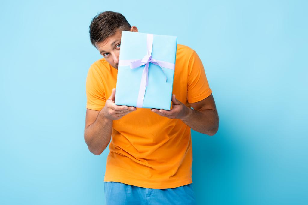 homme en t-shirt caché derrière boîte cadeau enveloppé sur bleu - Photo, image