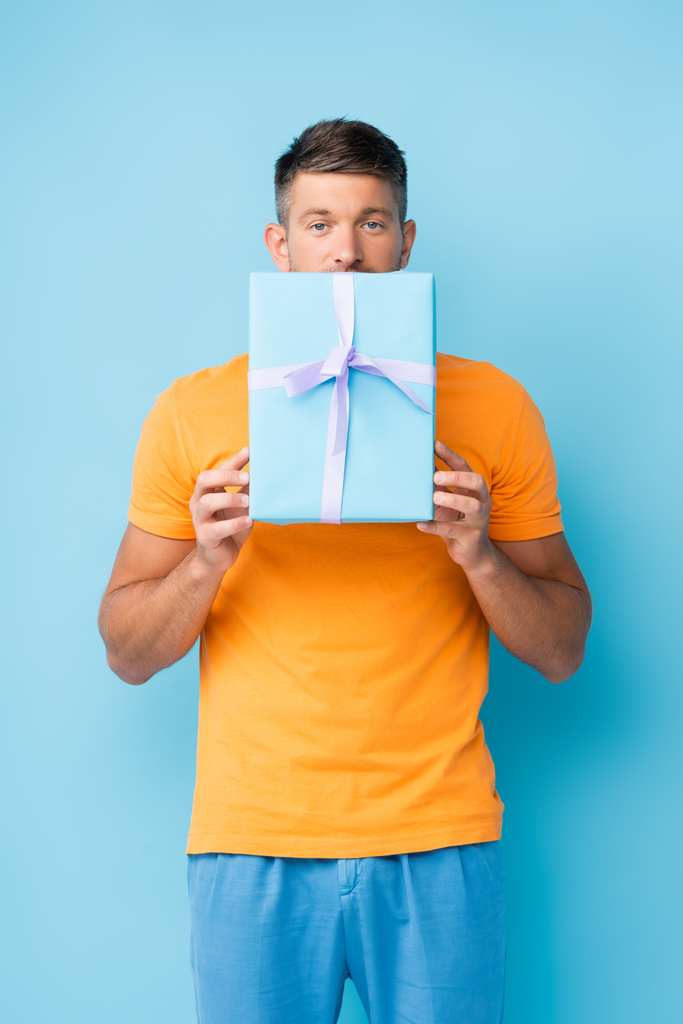 man in t-shirt covering mouth with wrapped gift box on blue - Photo, Image