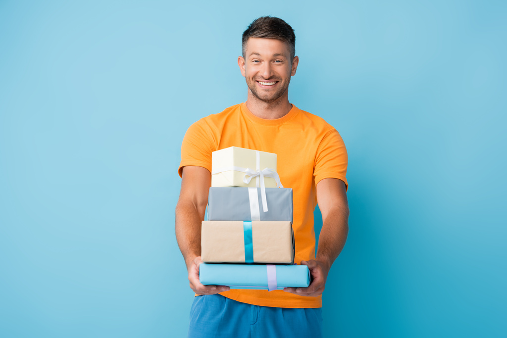 cheerful man in t-shirt holding wrapped presents on blue - Photo, Image