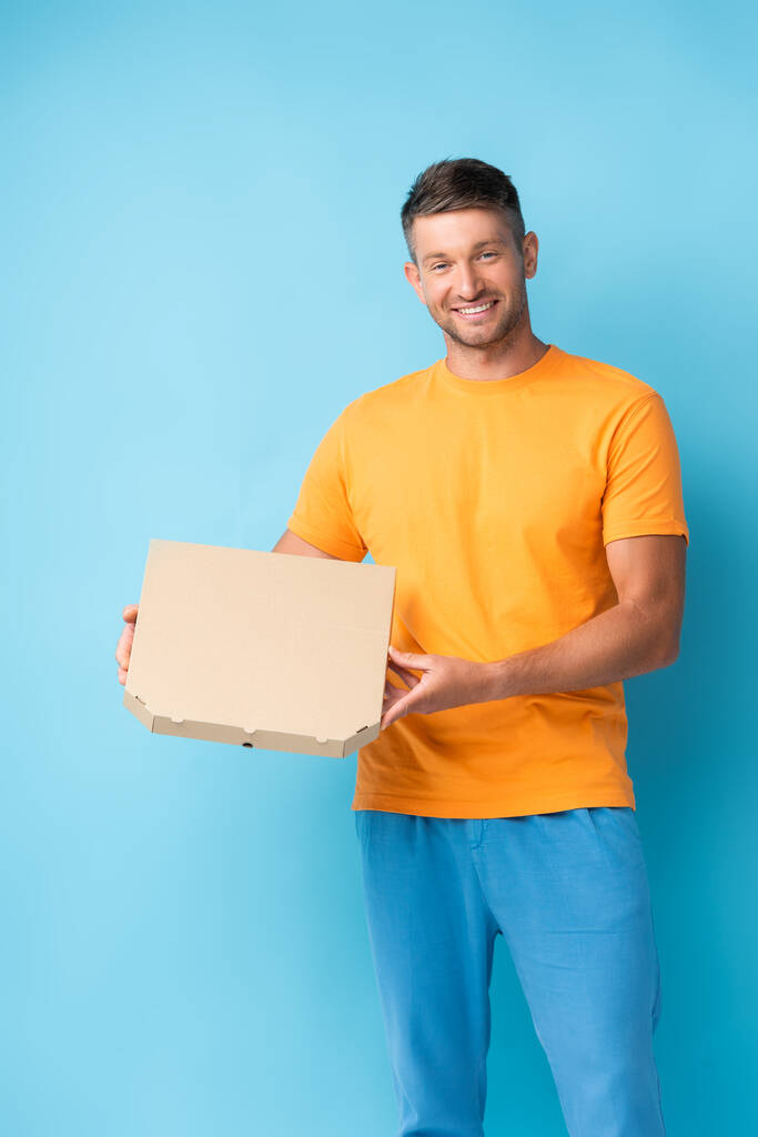 radostný muž v tričku drží kartónovou krabici od pizzy na modrém - Fotografie, Obrázek