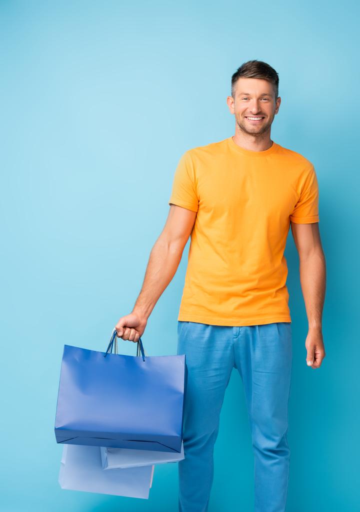 glimlachende man in t-shirt met boodschappentassen op blauw - Foto, afbeelding