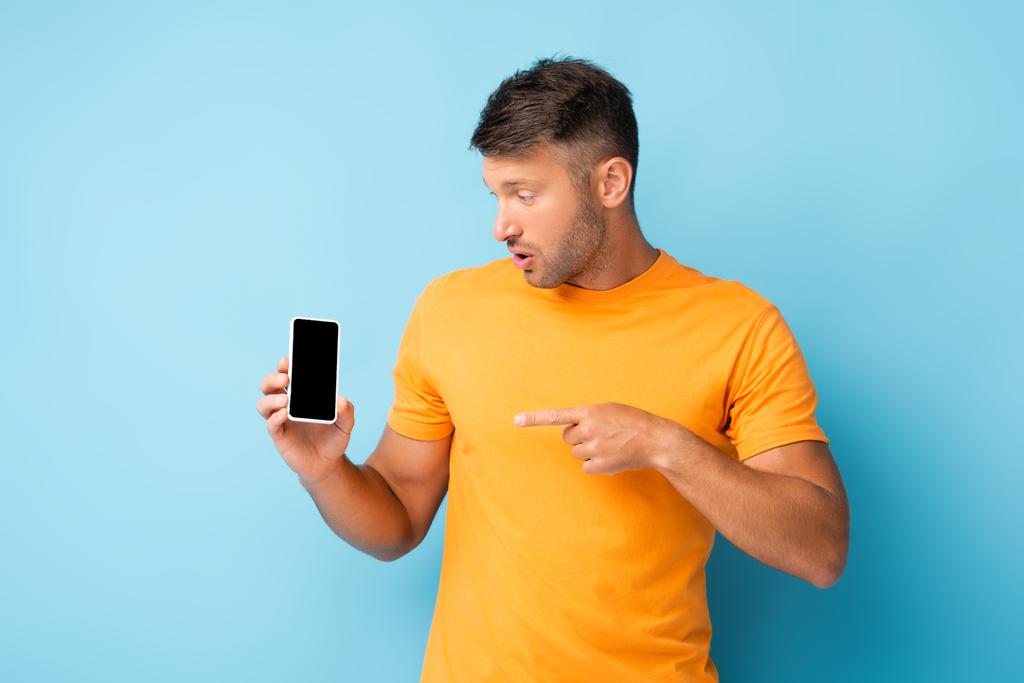 zaskoczony mężczyzna w t-shirt wskazując palcem na smartfona z pustym ekranem na niebiesko - Zdjęcie, obraz